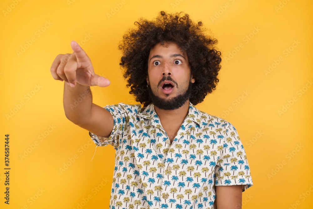 Φωτογραφία Young man with afro hair over wearing hawaiian shirt standing over yellow background Pointing with finger surprised ahead, open mouth amazed expression, something on the front