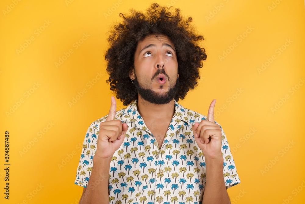 Φωτογραφία Young man with afro hair over wearing hawaiian shirt standing over yellow background amazed and surprised looking up and pointing with fingers and raised arms