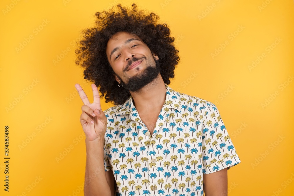 Φωτογραφία Young man with afro hair over wearing hawaiian shirt standing over yellow background smiling with happy face winking at the camera doing victory sign