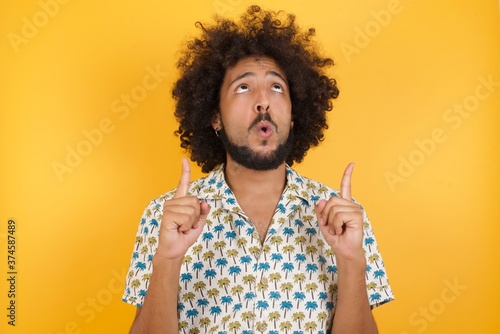Young man with afro hair over wearing hawaiian shirt standing over yellow background amazed and surprised looking up and pointing with fingers and raised arms.