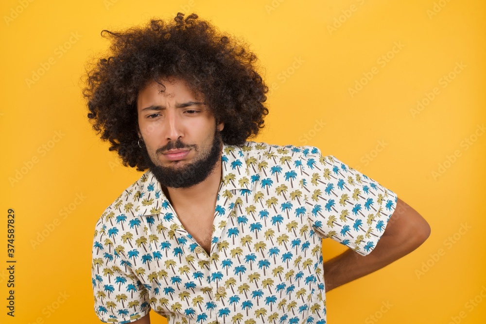 Young man with afro hair over wearing hawaiian shirt standing over yellow background Suffering of backache, touching back with hand, muscular pain Young man with afro hair over wearing hawaiian shirt standing over yellow background Suffering of backache, touching back with hand, muscular pain