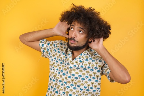 Young man with afro hair over wearing hawaiian shirt standing over yellow background Trying to hear both hands on ear gesture  curious for gossip. Hearing problem  deaf