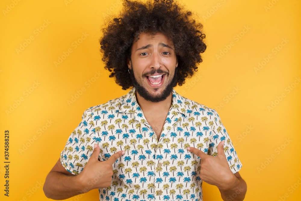 Horizontal shot of Young man with afro hair wearing hawaiian shirt stands indoors against concrete wall points at his body, being in good mood after going shopping and making successful purchases Horizontal shot of Young man with afro hair wearing hawaiian shirt stands indoors against concrete wall points at his body, being in good mood after going shopping and making successful purchases