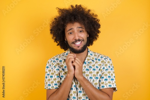 Positive adorable Young man with afro hair wearing hawaiian shirt standing over  smiles happily, glad to receive pleasant news from interlocutor, keeps hands together. People emotions concept.
