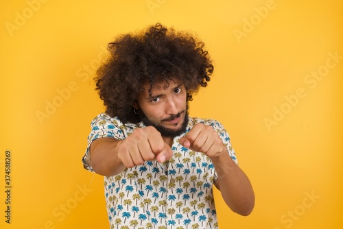 Portrait of strong and determined Young man with afro hair wearing hawaiian shirt standing over yellow wall punching air with fist and looking confidently at camera male struggle, fighting spirit © Roquillo