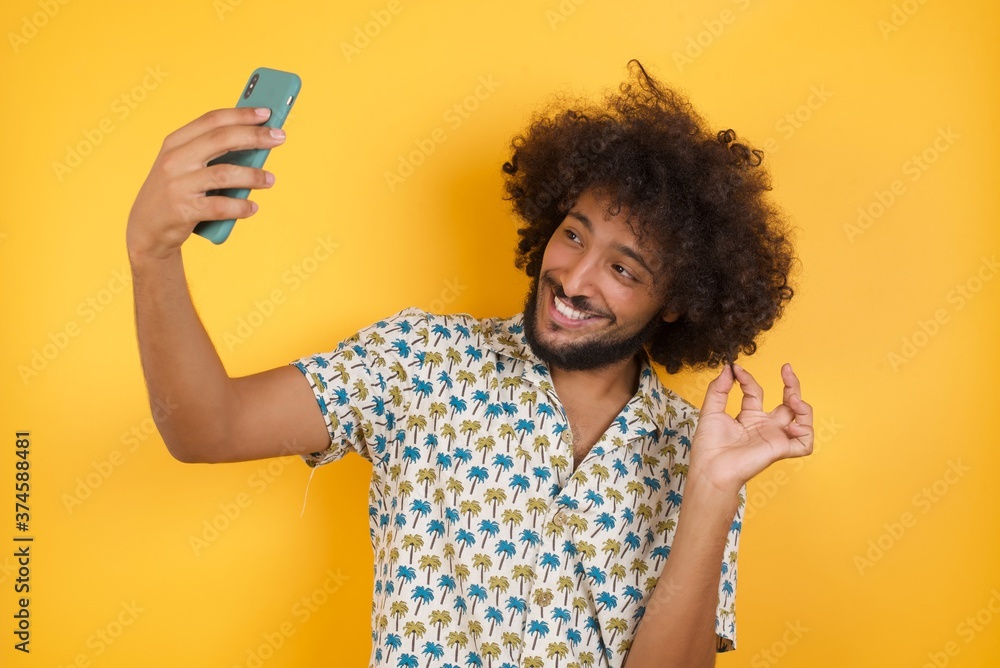 Photo of charming Young man with afro hair over wearing hawaiian shirt standing over yellow background smiling and taking a selfie ready to post it on her social media. Photo of charming Young man with afro hair over wearing hawaiian shirt standing over yellow background smiling and taking a selfie ready to post it on her social media.