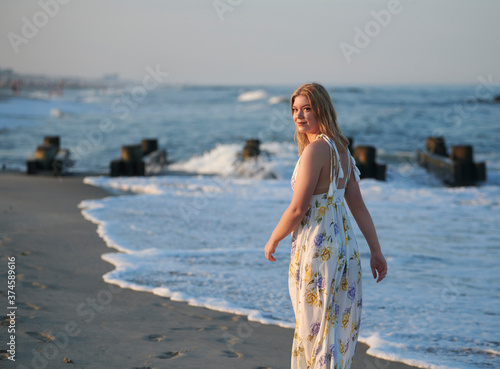Pretty young blonde woman in dress walks along the water at beach - summer vacation