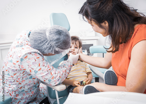 Cute caucasian baby boy sitting on a kids dentist chair having his mouth checked by a female dentist in her clinic and his mother comforting him. Doctor is using protective hair cap and face shield. photo