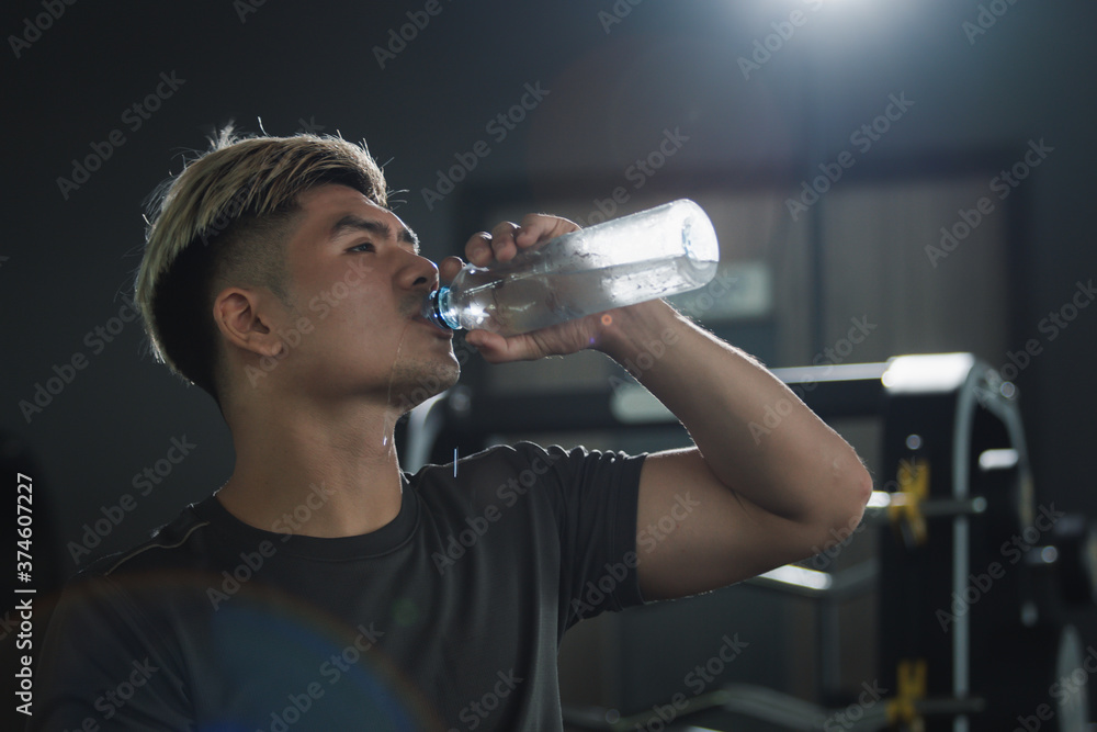 Strong sportive man relaxing and drinking water from a water bottle after exercised in a indoor gym. Young Asian Athletes refresh himself after workout by drinking a cold water. Image with copyspace. Strong sportive man relaxing and drinking water from a water bottle after exercised in a indoor gym. Young Asian Athletes refresh himself after workout by drinking a cold water. Image with copyspace.