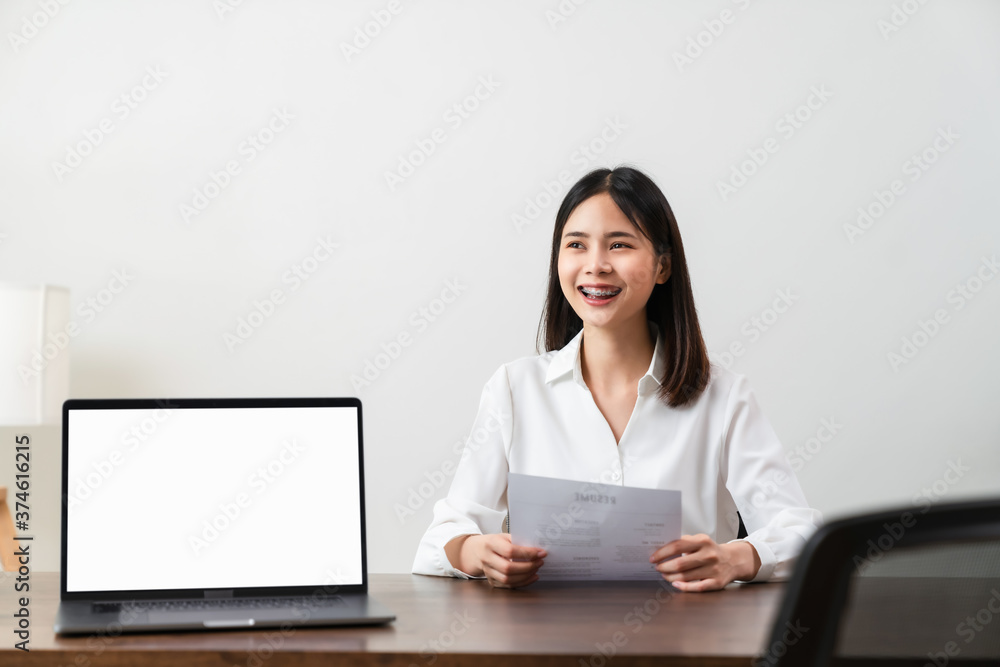 Smiley young Asian woman sitting at a wooden table. Hold resume application information and wait for interviews from company personnel. The digital laptop screen is blank on the desk. Smiley young Asian woman sitting at a wooden table. Hold resume application information and wait for interviews from company personnel. The digital laptop screen is blank on the desk.