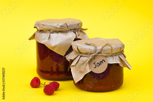 Raspberry jam in a glass jar and fresh raspberry on yellow background.