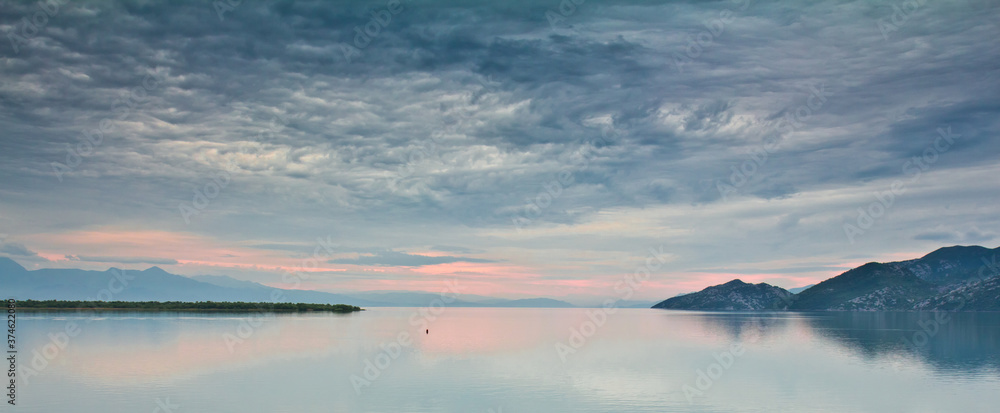 Skadar lake at sunrise in Montenegro. Smooth surface of water with reflection of mountains against cloudy colorful sky.