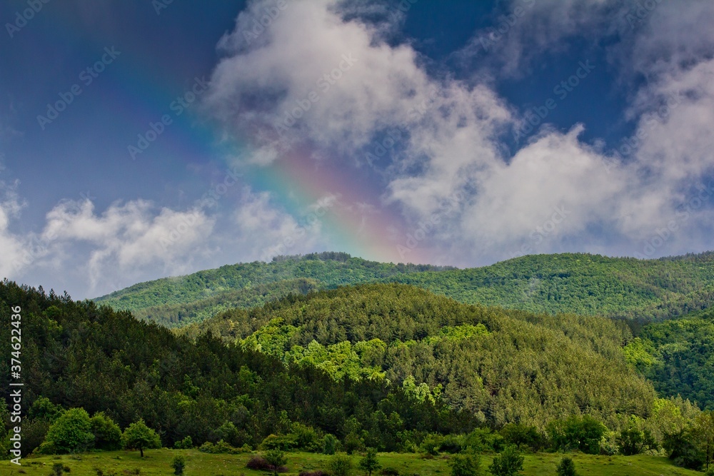 Rainbow above green field and forest on hills of Rhodope mountains in a sunny day in Bulgaria.