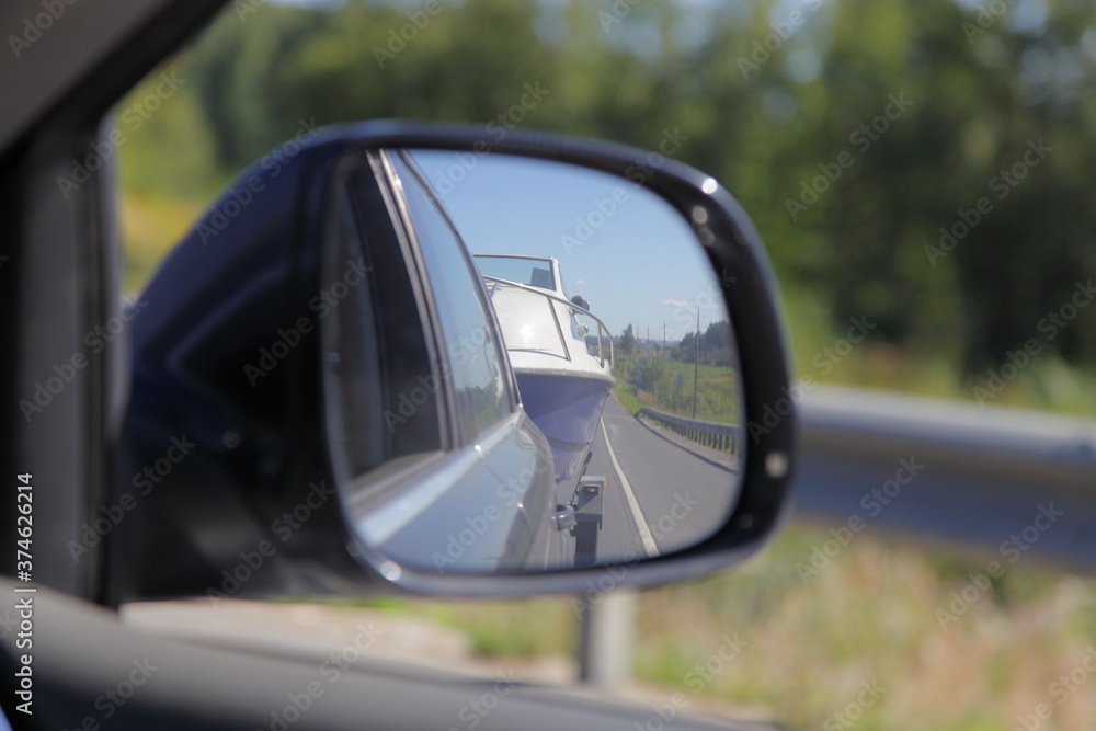 Close up view on reflection in right side car rear mirrow to towing motor boat on car trailer on blurred roadside fencing and green bushes background, road trip with private boat