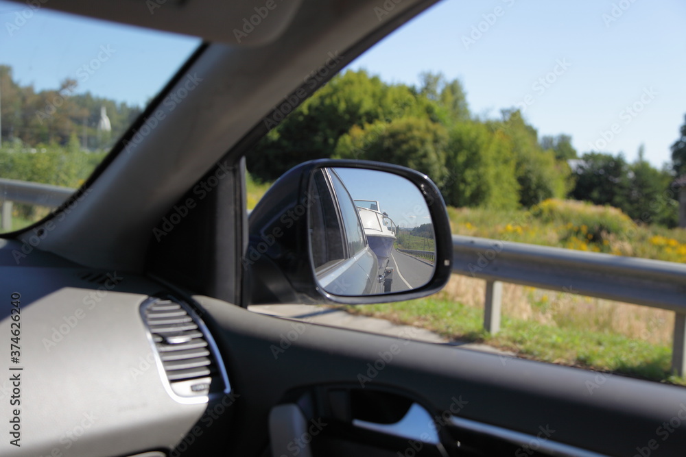 Indoor to outdoor from car view in front side window to back view mirror with boat on trailer and natural landscape reflection, car trip active vacation in Russia on Sunny summer day