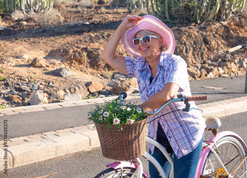 Obraz premium Cheerful senior woman with pink hat and sunglasses shows the tongue riding with vintage bicycle enjoying freedom, basket with daisies - concept of active playful elderly during vacation