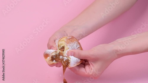 Hands tear open a fresh round stuffed donut. Condensed milk is pouring inside. Lots of powdered sugar. Pink background. Delicious food.