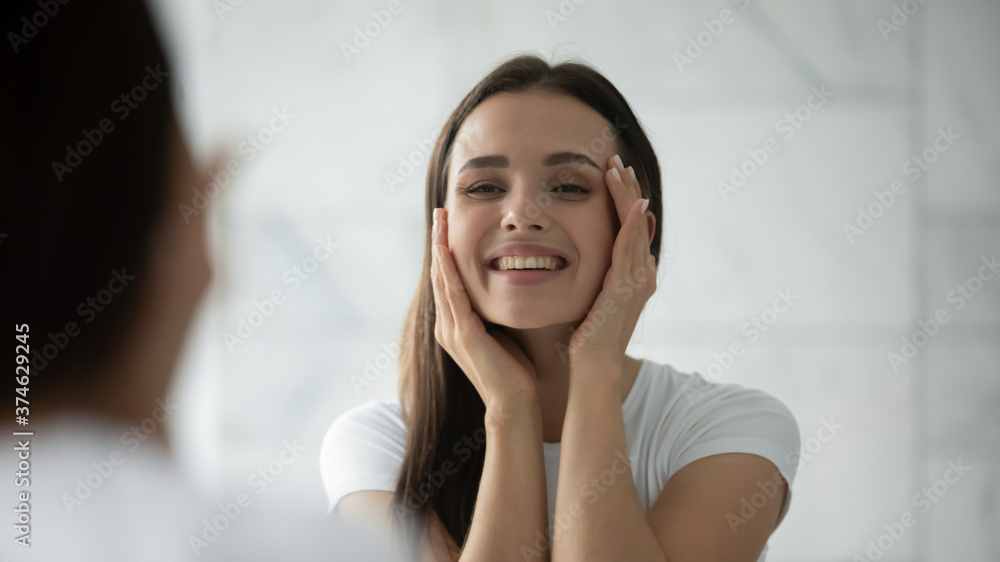 Close up head shot smiling satisfied young woman touching face, standing in bathroom, looking at mirror, happy female wearing white t-shirt doing facial massage, enjoying perfect smooth skin Close up head shot smiling satisfied young woman touching face, standing in bathroom, looking at mirror, happy female wearing white t-shirt doing facial massage, enjoying perfect smooth skin