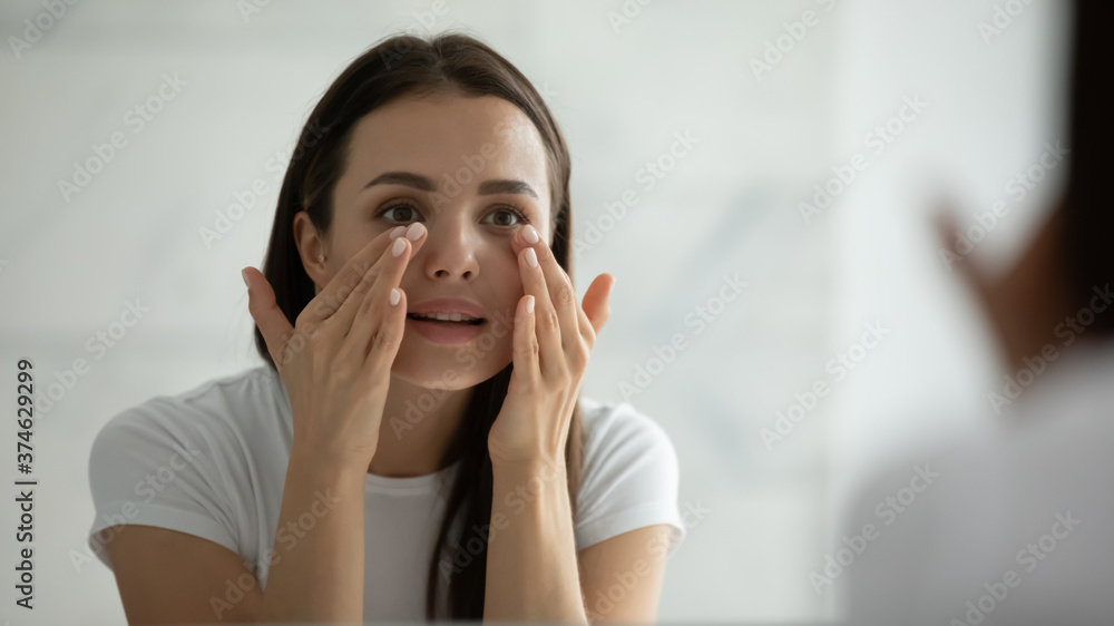 Close up smiling young woman wearing white t-shirt doing facial massage, applying moisturizing cream on under eye skin, looking in mirror, standing in bathroom, enjoying skincare procedure Close up smiling young woman wearing white t-shirt doing facial massage, applying moisturizing cream on under eye skin, looking in mirror, standing in bathroom, enjoying skincare procedure