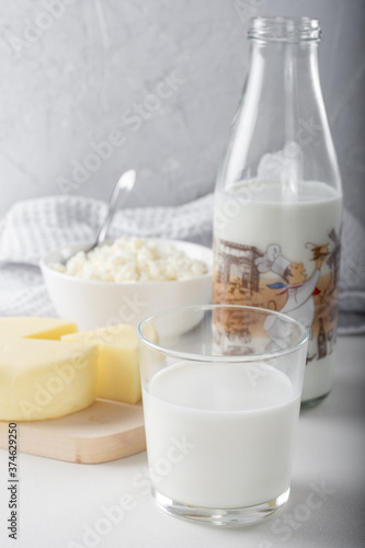 A glass of milk with a bottle of milk, cottage cheese and a circle of cheese in the background, on a light background.