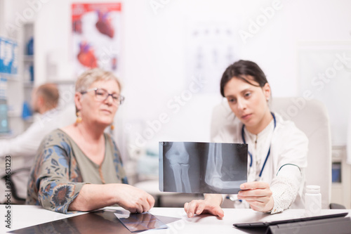 Doctor discussing with senior patient about her leg injury while holding x-ray in hospital office. Medic wearing white coat working on computer.