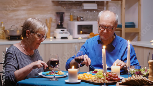 Cheerful old retired couple in love having meal at home. Aged mature couple eating and discussing during romantic dinner sitting at the table in the modern kitchen, enjoying time together
