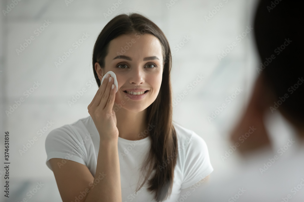 Close up head shot smiling young woman wearing white t-shirt removing face makeup, using cotton pad, standing in bathroom, looking in mirror, cleaning face, enjoying skincare procedure Close up head shot smiling young woman wearing white t-shirt removing face makeup, using cotton pad, standing in bathroom, looking in mirror, cleaning face, enjoying skincare procedure