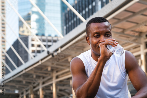 portrait of refreshing young african american sportsman drinking water from bottle after running exercise in city in morning. sports exercise and city lifestyle.