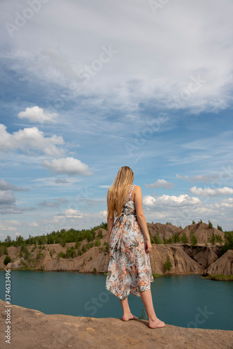 A girl with long blond hair in a light dress and bare feet stands on a cliff above the river.