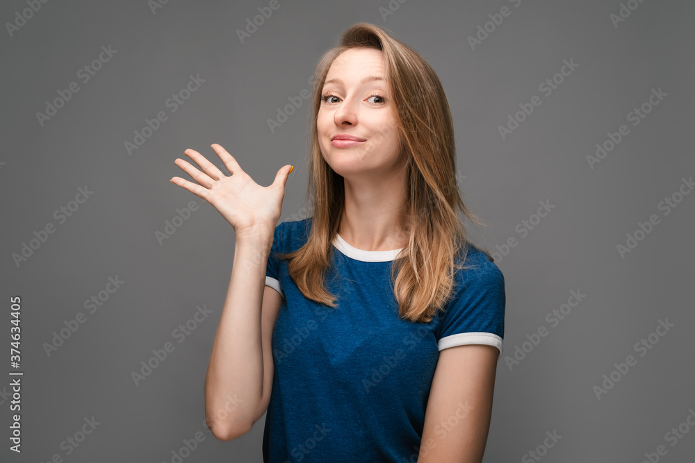 Positive Young female with blonde straight hair, dressed in blue t shirt, smiling and spreads her hand to the side Positive Young female with blonde straight hair, dressed in blue t shirt, smiling and spreads her hand to the side