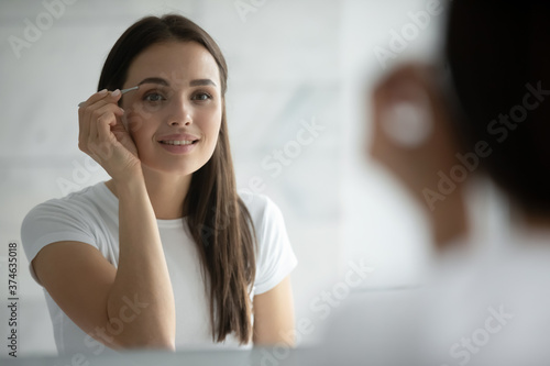 Close up head shot smiling young woman wearing white t-shirt plucking eyebrows with metal tweezers, doing correcting shape procedure, standing in bathroom at home, looking in mirror