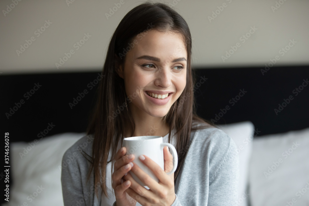 Close up head shot smiling young woman holding white cup of tea or coffee, sitting on bed in bedroom at home, enjoying breakfast in morning, happy overjoyed beautiful girl drinking hot beverage Close up head shot smiling young woman holding white cup of tea or coffee, sitting on bed in bedroom at home, enjoying breakfast in morning, happy overjoyed beautiful girl drinking hot beverage