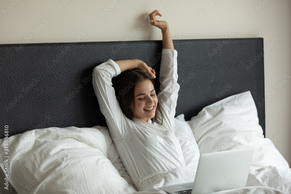Smiling beautiful young woman stretching hands in cozy bed, using laptop, watching movie, chatting on social network, working online at home, relaxing with computer, looking at screen Smiling beautiful young woman stretching hands in cozy bed, using laptop, watching movie, chatting on social network, working online at home, relaxing with computer, looking at screen