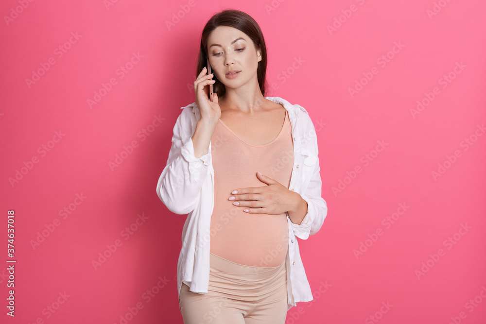 Pregnant woman in kitchen talking on cellular phone with serious and concentrated facial expression, wearing stylish leggins and bodysuit, touching her stomach. Pregnant woman in kitchen talking on cellular phone with serious and concentrated facial expression, wearing stylish leggins and bodysuit, touching her stomach.