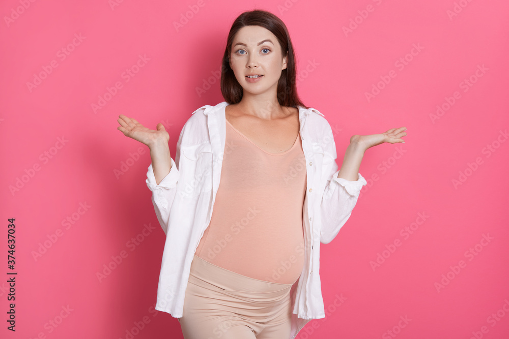 Beautiful dark haired european pregnant woman in comfy outfit spreading hands with astonish expression, doesn't know gender of her future baby, posing against pink wall. Beautiful dark haired european pregnant woman in comfy outfit spreading hands with astonish expression, doesn't know gender of her future baby, posing against pink wall.