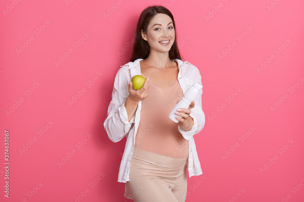 Beautiful pregnant woman with green apple and bottle of milk in hands, looking directly at camera while posing against pink wall, healthy eating for expectant mother. Beautiful pregnant woman with green apple and bottle of milk in hands, looking directly at camera while posing against pink wall, healthy eating for expectant mother.