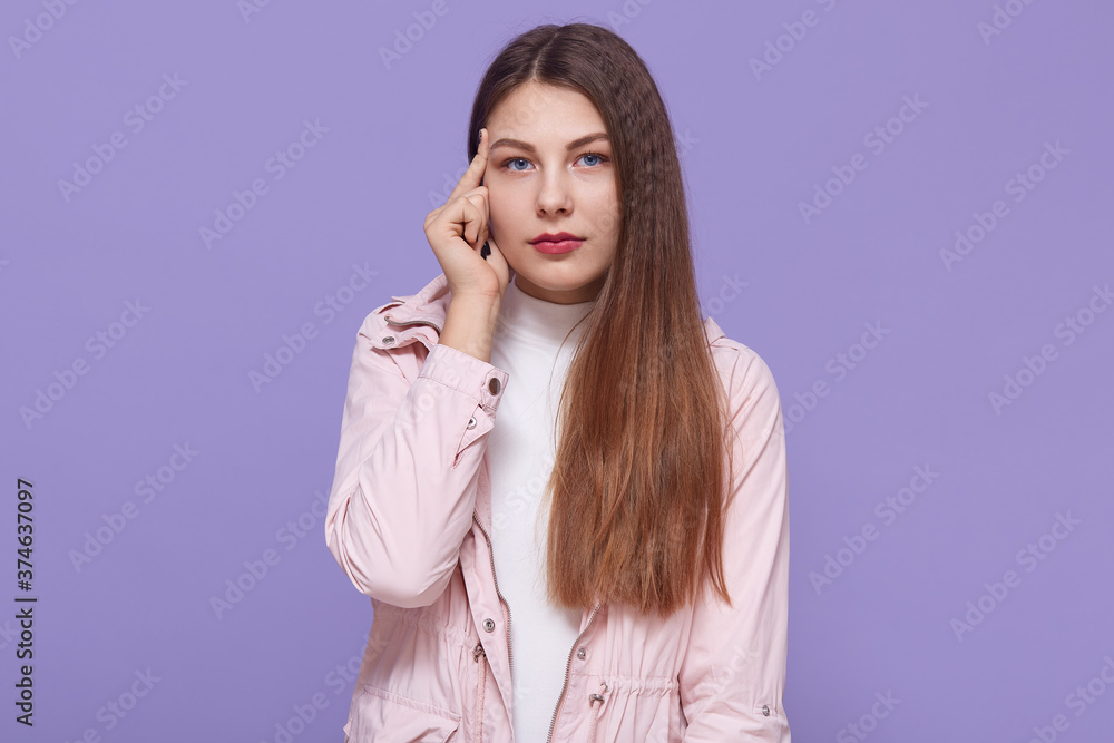 Fototapeta premium Young thoughtful female has serious pensive expression, keeping finger near temple, wearing pale pink jacket, stands against lilac background, looks at camera.