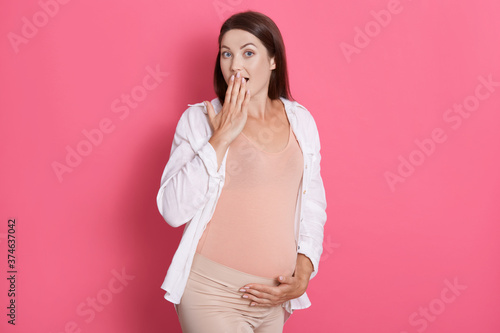 Surprised pregnant woman posing with opened mouth  covering mouth with palm  posing isolated over pink background  touching her belly  wearing leggins and shirt.