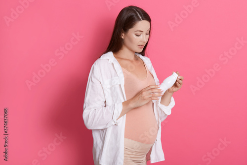 Fotografie Attractive pregnant woman reading composition of product, holding bottle or yogurt or milk, standing against pink wall, healthy eating for future mother and baby