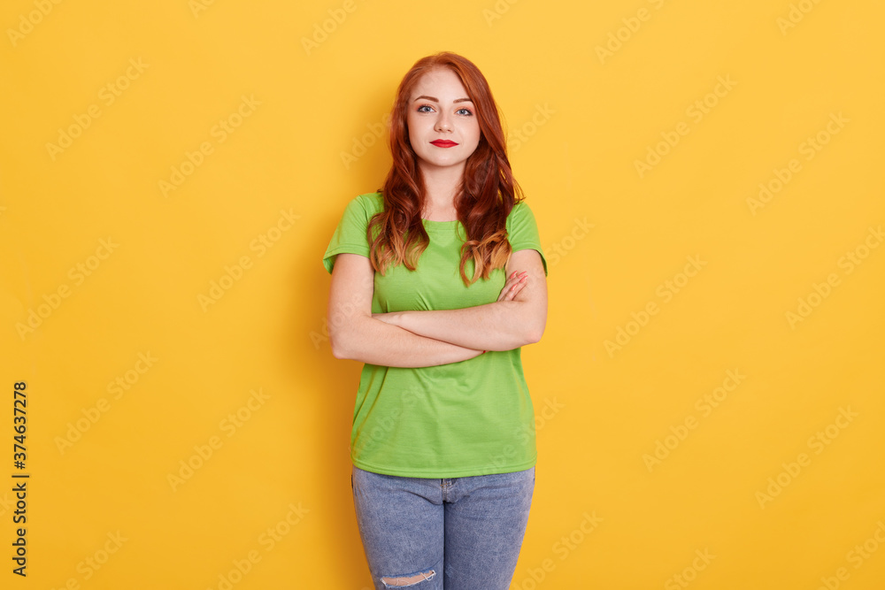 Cute girl in casual green t shirt and standing with arms folded against yellow background, being serious, looking at camera, has red wavy hair and bright lips. Cute girl in casual green t shirt and standing with arms folded against yellow background, being serious, looking at camera, has red wavy hair and bright lips.