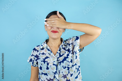 Young beautiful woman standing over blue isolated background serious and covering her eyes with her hand.
