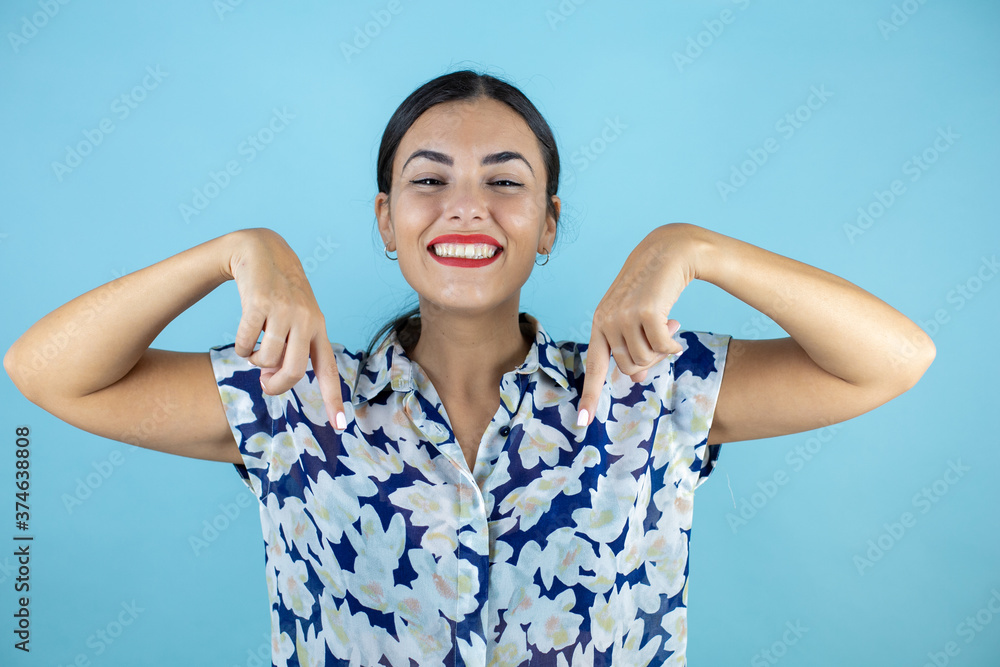 Young beautiful woman wearing sunglasses over isolated blue background smiling, looking and pointing down with fingers and raised arms. Young beautiful woman wearing sunglasses over isolated blue background smiling, looking and pointing down with fingers and raised arms.