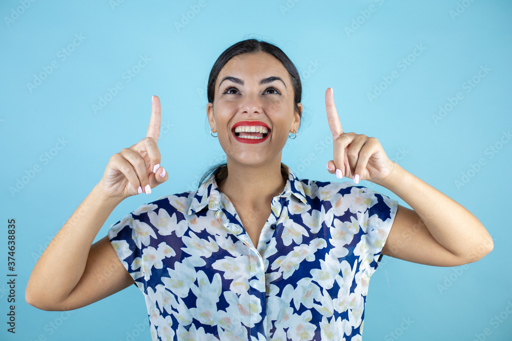 Young beautiful woman wearing sunglasses over isolated blue background smiling, looking and pointing up with fingers and raised arms.