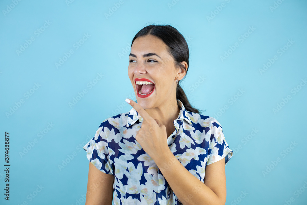 Young beautiful woman standing over isolated blue background smiling looking to the side and pointing to the empty space with one finger. Young beautiful woman standing over isolated blue background smiling looking to the side and pointing to the empty space with one finger.