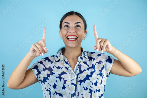 Young beautiful woman wearing sunglasses over isolated blue background smiling, looking and pointing up with fingers and raised arms.