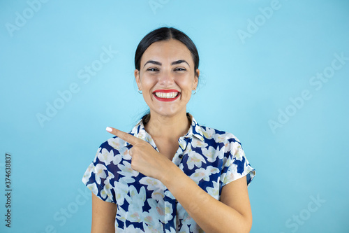 Young beautiful woman standing over isolated blue background smiling looking to the camera and pointing to the empty space with one finger.