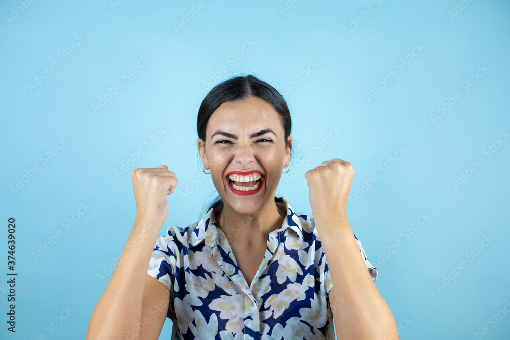 Young beautiful woman standing over isolated blue background screaming with her fists in the air with a gesture of victory. Young beautiful woman standing over isolated blue background screaming with her fists in the air with a gesture of victory.
