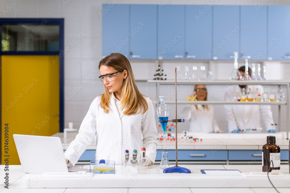 Female researcher in white lab coat and protective mask using laptop while working in the lab Female researcher in white lab coat and protective mask using laptop while working in the lab