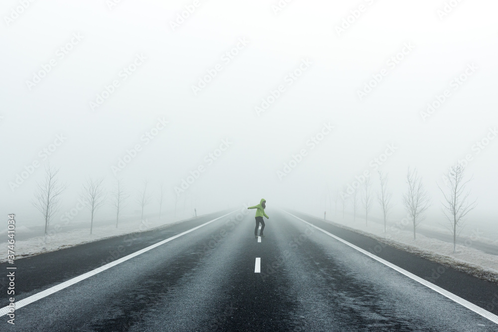 Woman in a green jacket walking down the middle of the road into the thick fog while looking back.