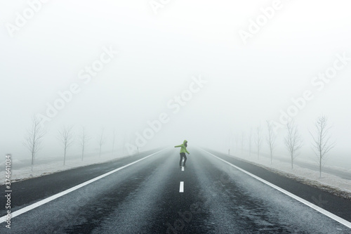 Woman in a green jacket walking down the middle of the road into the thick fog while looking back.